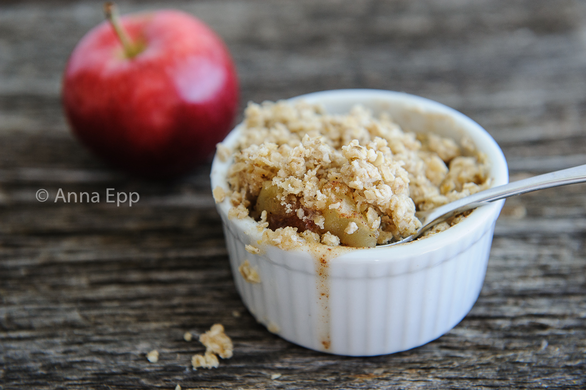 Apple crisp in a ramkin with a spoon, red apple in the background