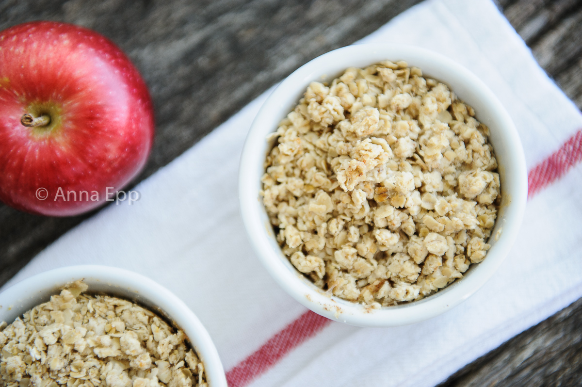 two baked apple crisps in white ramkins sitting on a white napkin with red ribbing, a red apple sits off to the side